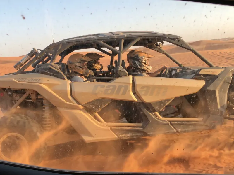 Four-seat Can-Am Maverick buggy throwing up sand as it crosses the dunes, seen through a vehicle window