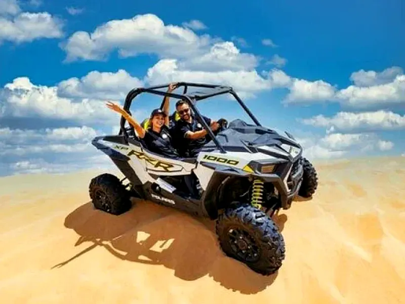 Couple waving from a white 2-seat Polaris RZR buggy on top of a red sand dune