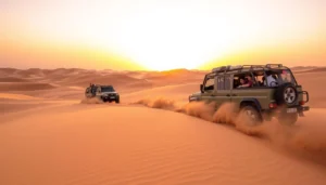 Two 4x4 safari vehicles with passengers driving over desert dunes at sunset, kicking up trails of sand