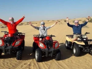 Tourists riding quad bikes across golden sand dunes in the Dubai desert at sunset, kicking up sand as they navigate the terrain