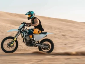 Tourists riding dirt bikes across the vast, sandy Dubai desert under a clear blue sky, kicking up sand as they go
