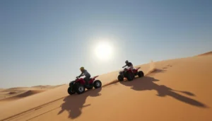 Two riders on red quad bikes climbing a sand dune under a bright sun in the Dubai desert